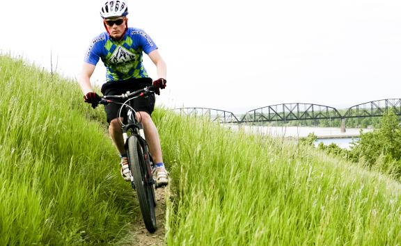 A mountain biker rides through tall green grass on a hillside trail