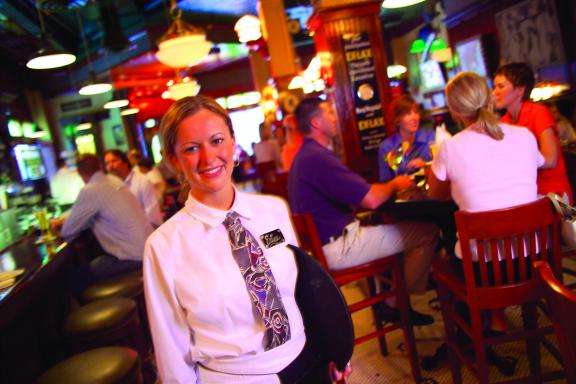 A smiling server stands in a busy bar or restaurant, wearing a white shirt and a patterned tie